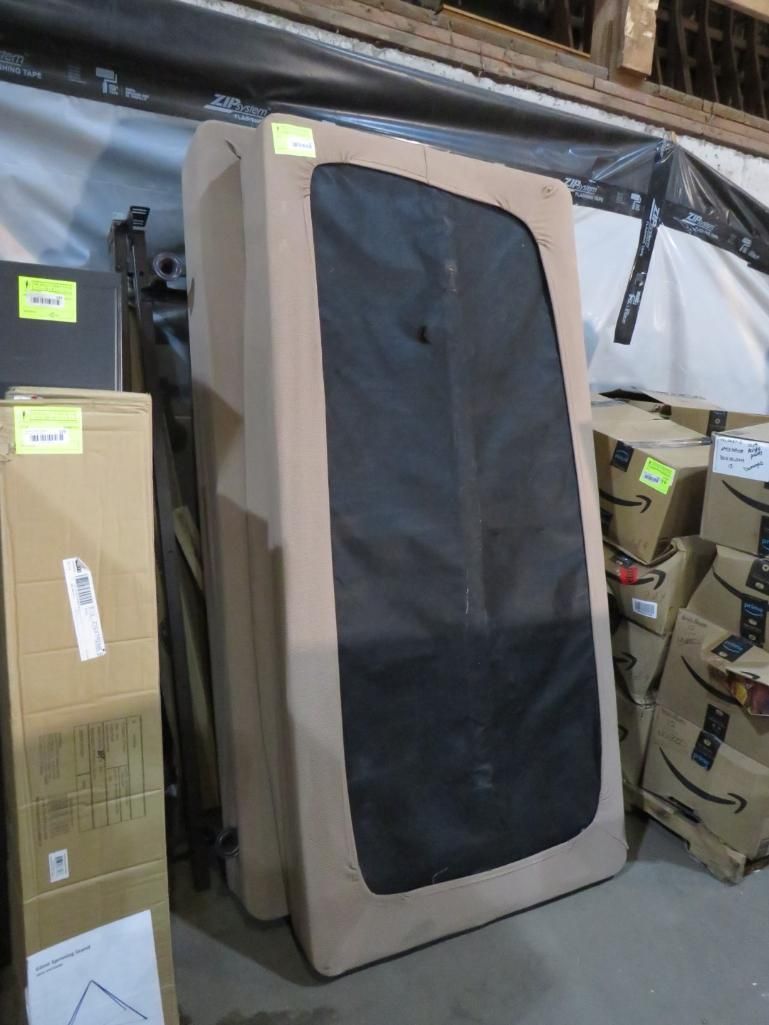 Tan upholstered mattress leaning, black mesh panel. Boxes stacked nearby in a warehouse.