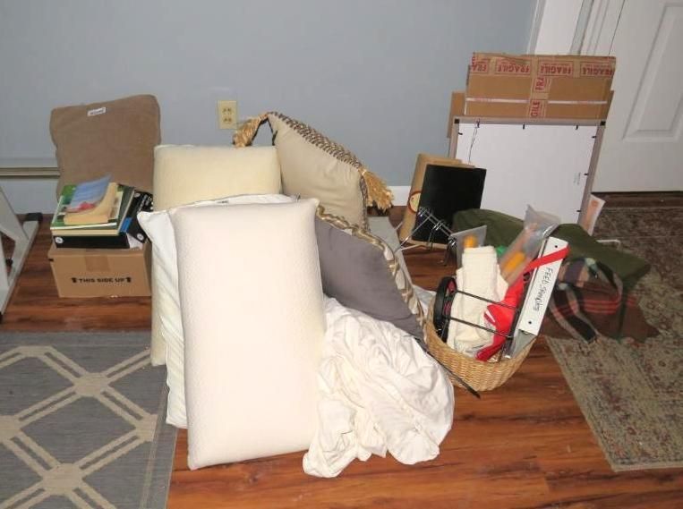 Pile of pillows, blankets, a basket, and boxes on a wooden floor against a blue wall.