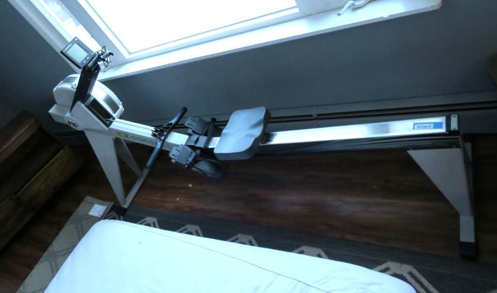 Rowing machine indoors, positioned near a window. Features a digital monitor and sliding seat on a track.