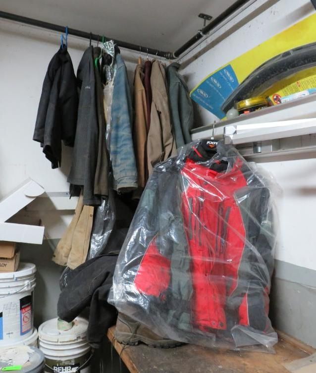 Coats hanging on a rod in a corner of a room, including a red and black jacket in a plastic bag.