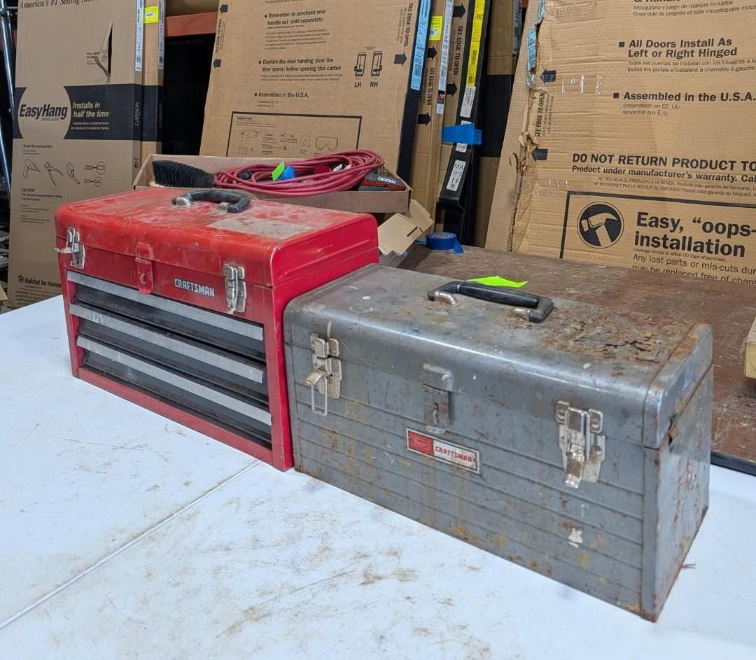 Red and gray metal toolboxes on a white table in a storage area with stacked boxes behind.