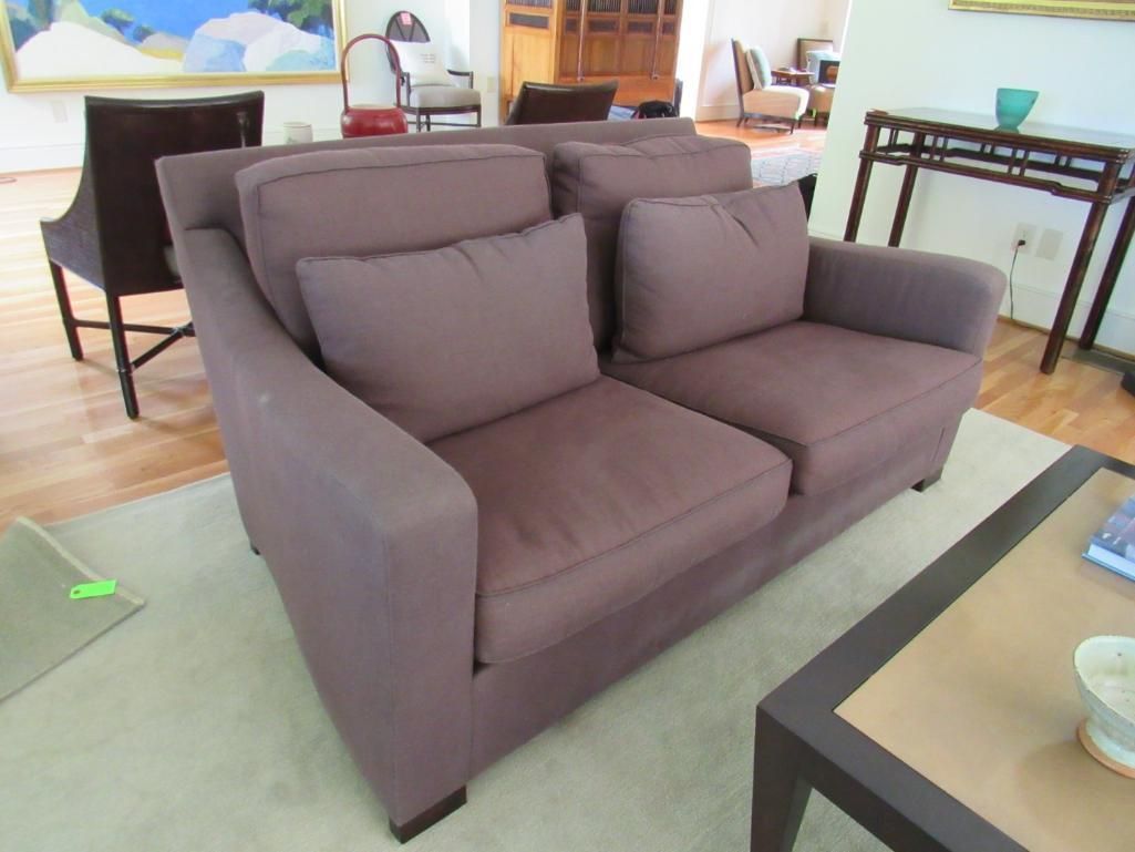 Mauve two-seater sofa with two cushions, set in a room with a rug and other furniture.