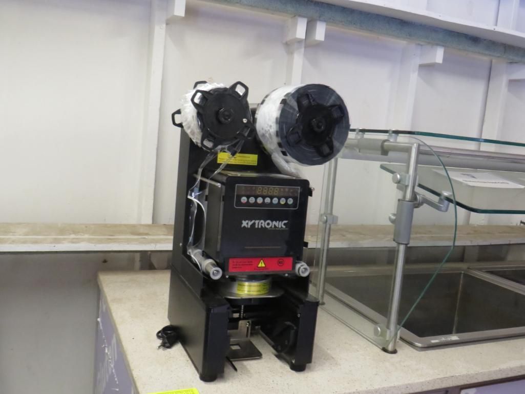 Black industrial machine with spools and digital display on a white surface next to a metal structure.