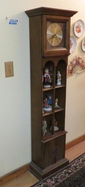 Tall wooden clock/display cabinet; clock face at the top, figurines on shelves below, with a cabinet at the base.