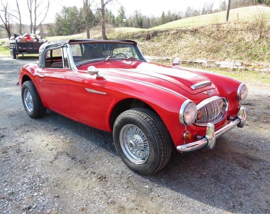 Red classic Austin-Healey convertible on a road, sunny outdoor setting.