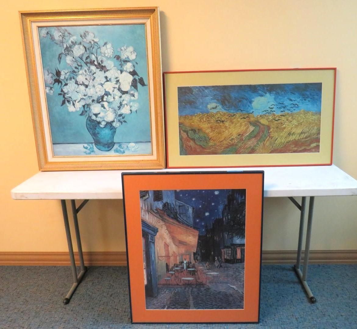 Three framed paintings on a white folding table: floral still life, wheat field, and a cafe scene.