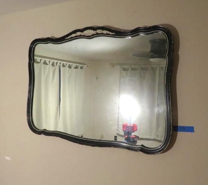 Rectangular ornate mirror with black frame reflecting a room with curtains and a lamp.