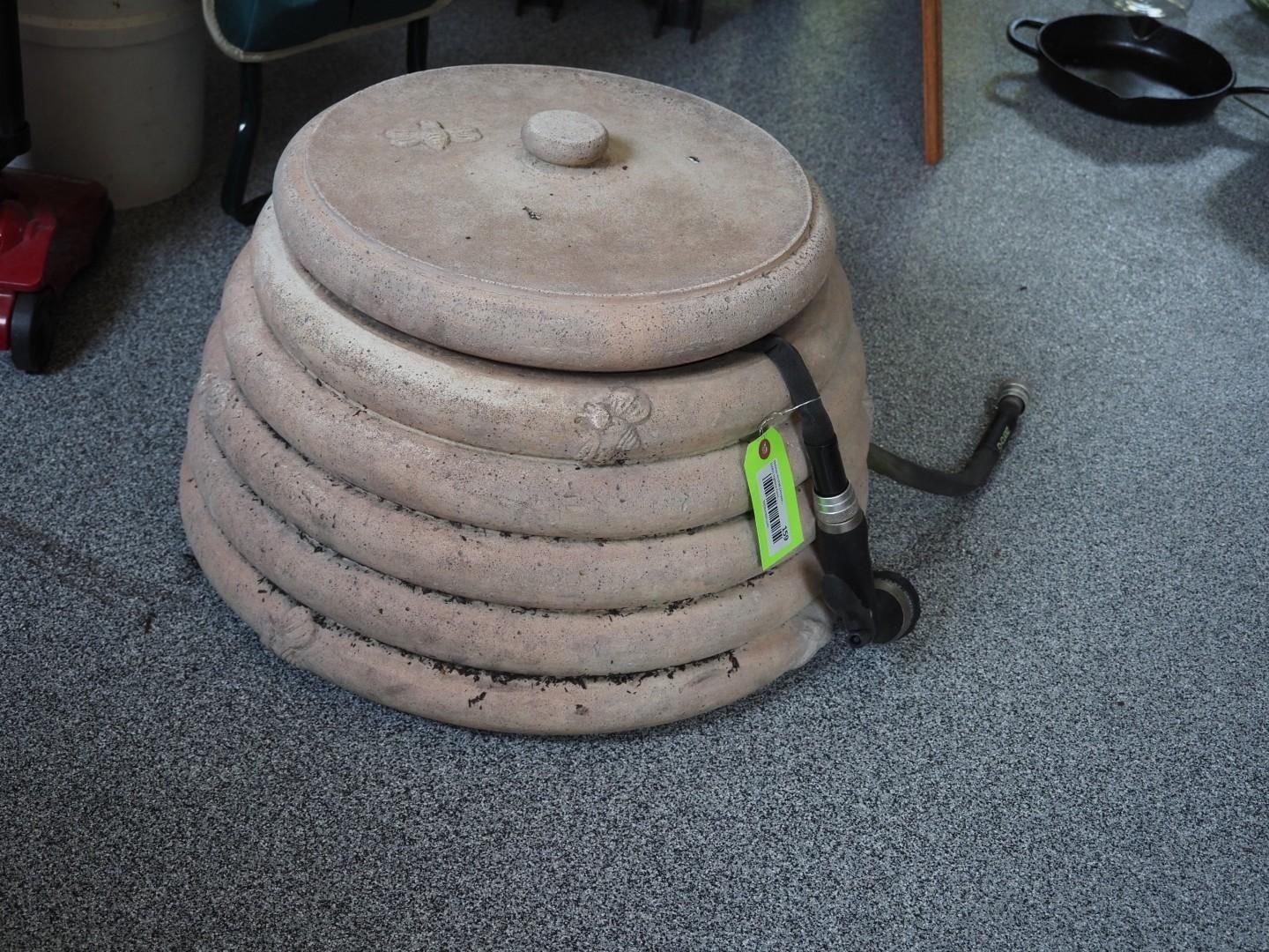 Round, beige outdoor water feature with lid, black hose attached, on a gray floor.