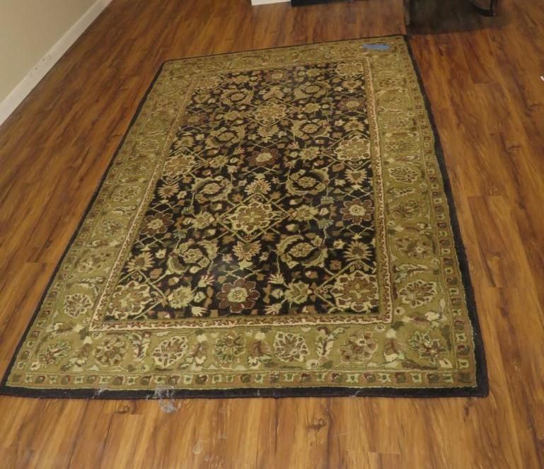An ornate, rectangular rug on a wood floor. The rug features dark, floral patterns with a green border.