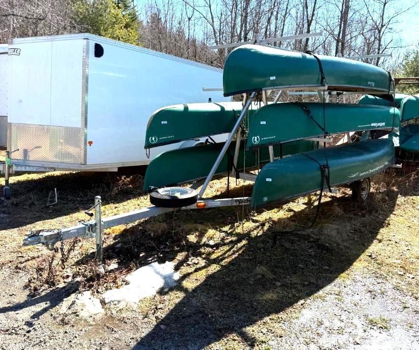 A trailer holding several green canoes.  White trailer is in the background. Sunny outdoor setting.