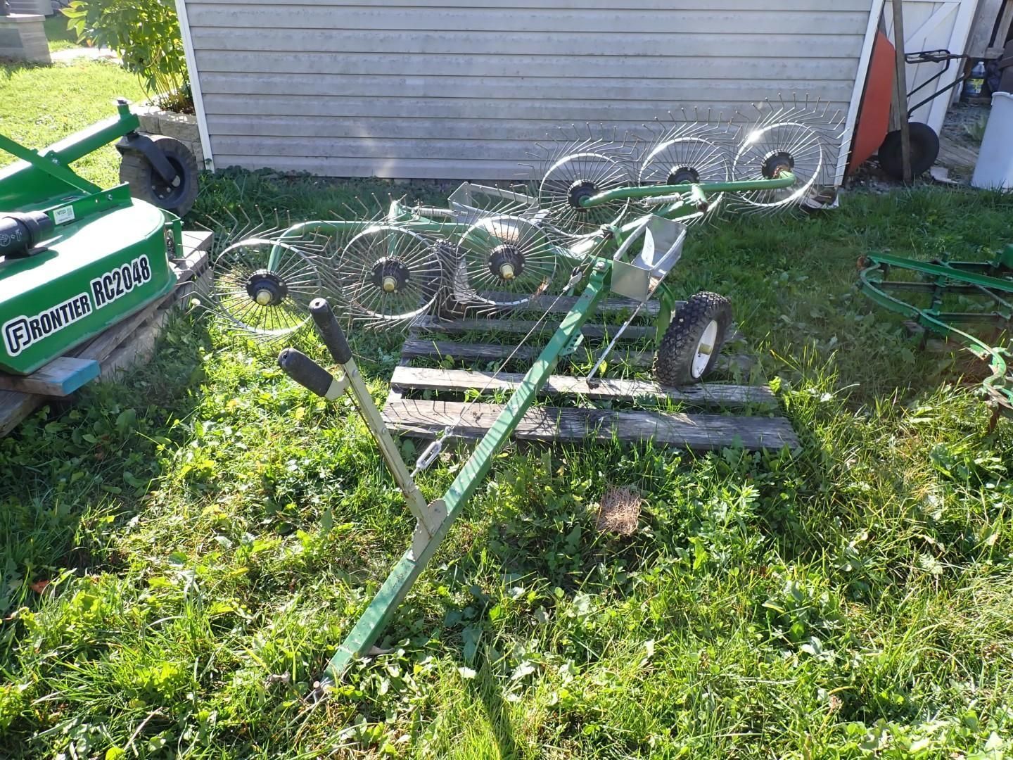 Green farm implement with rotating tines lying on grass beside a building.