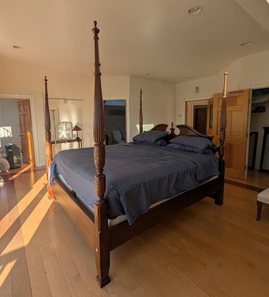 Spacious bedroom with a four-poster bed, dark blue bedding, wood floors, and warm sunlight.