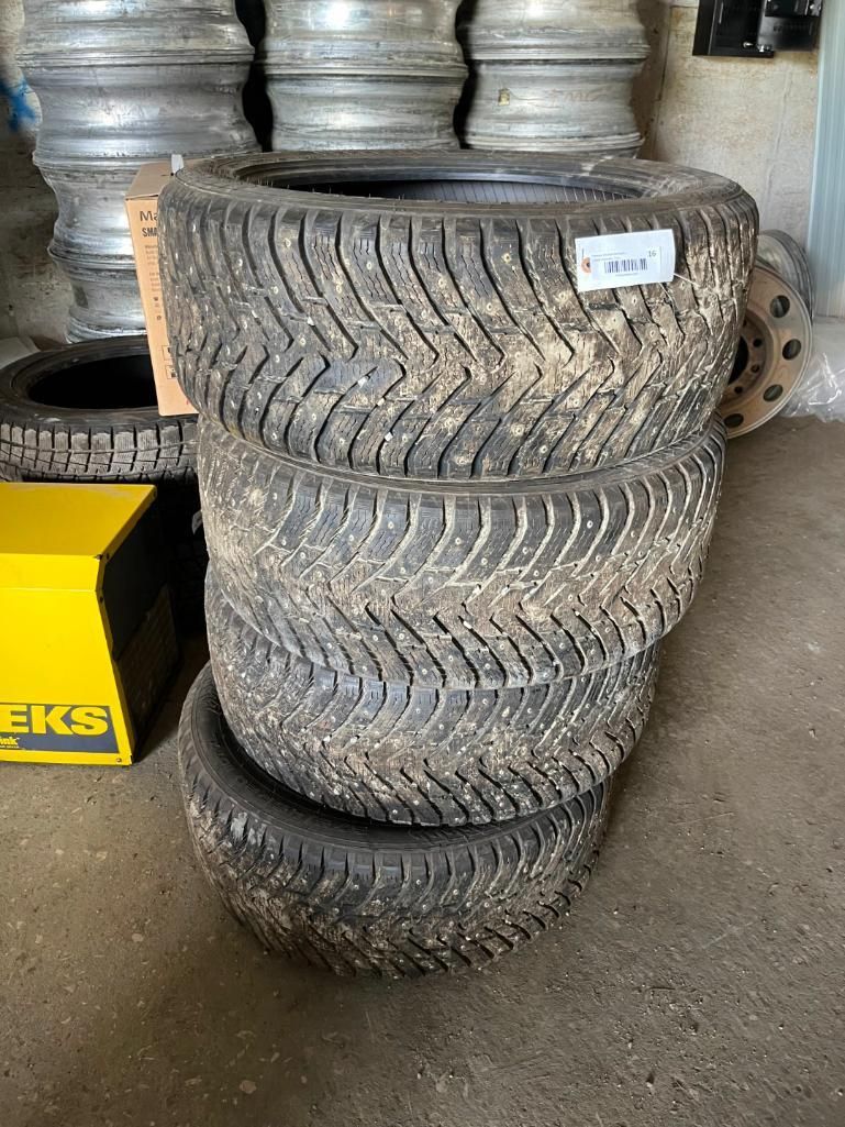 Four dirty, stacked tires with a mud-like tread pattern, with a stack of rims in the background.