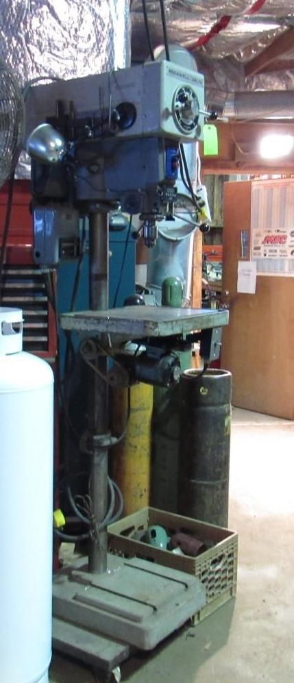 A large, industrial drill press in a workshop. It is light blue and gray, with a work table.