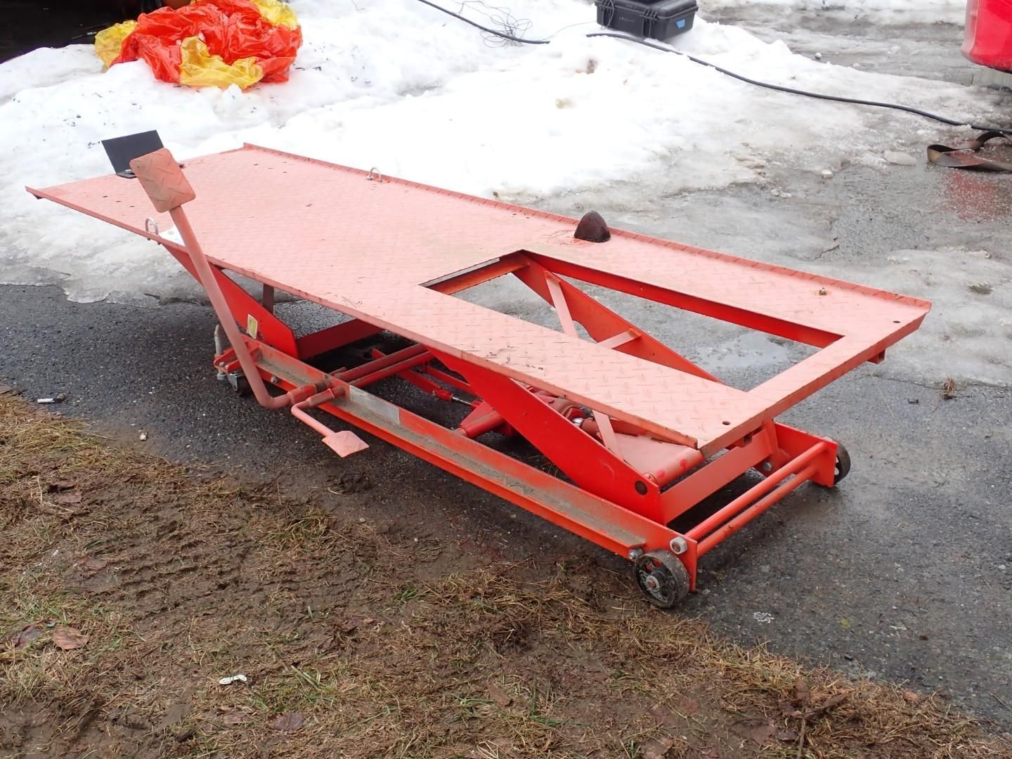 An orange hydraulic motorcycle lift table sits on a paved surface outdoors with snow in the background.