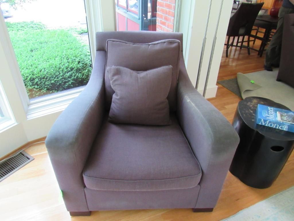Purple upholstered armchair with matching pillows, beside a window and small table.