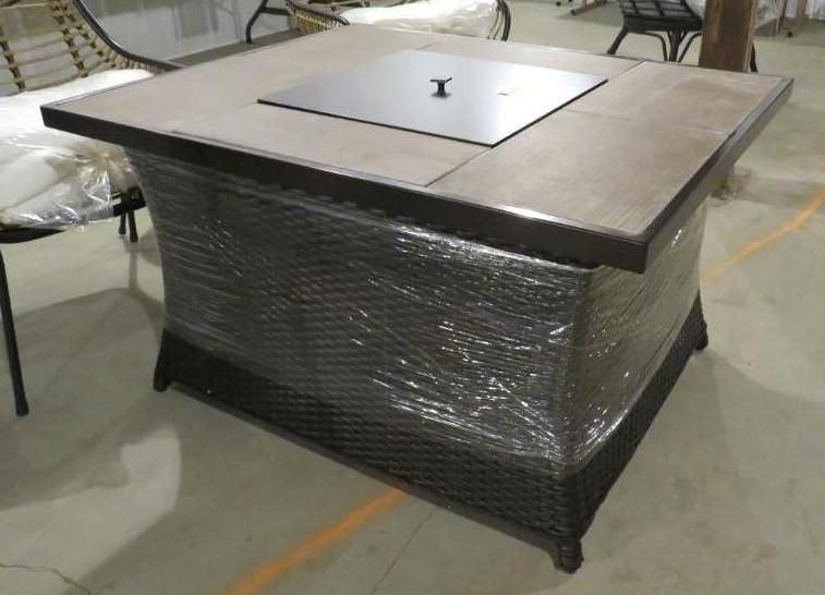 Square fire pit table with a stone top and woven base, wrapped in plastic.