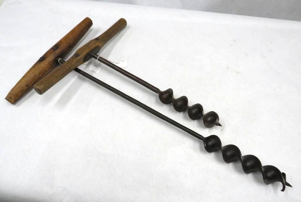 Two wooden-handled hand augers lie on a white surface. The augers have metal bits with spiral cutting edges.