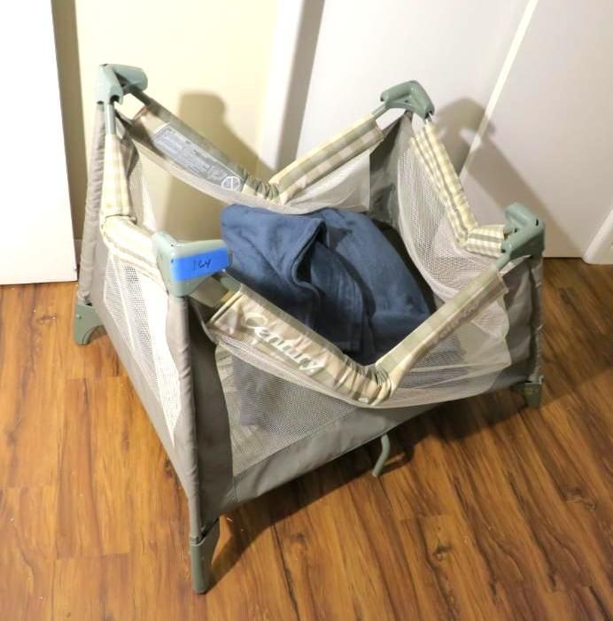 A small, open playard with beige fabric and mesh sides, containing a dark blue garment, sits on a wood floor.