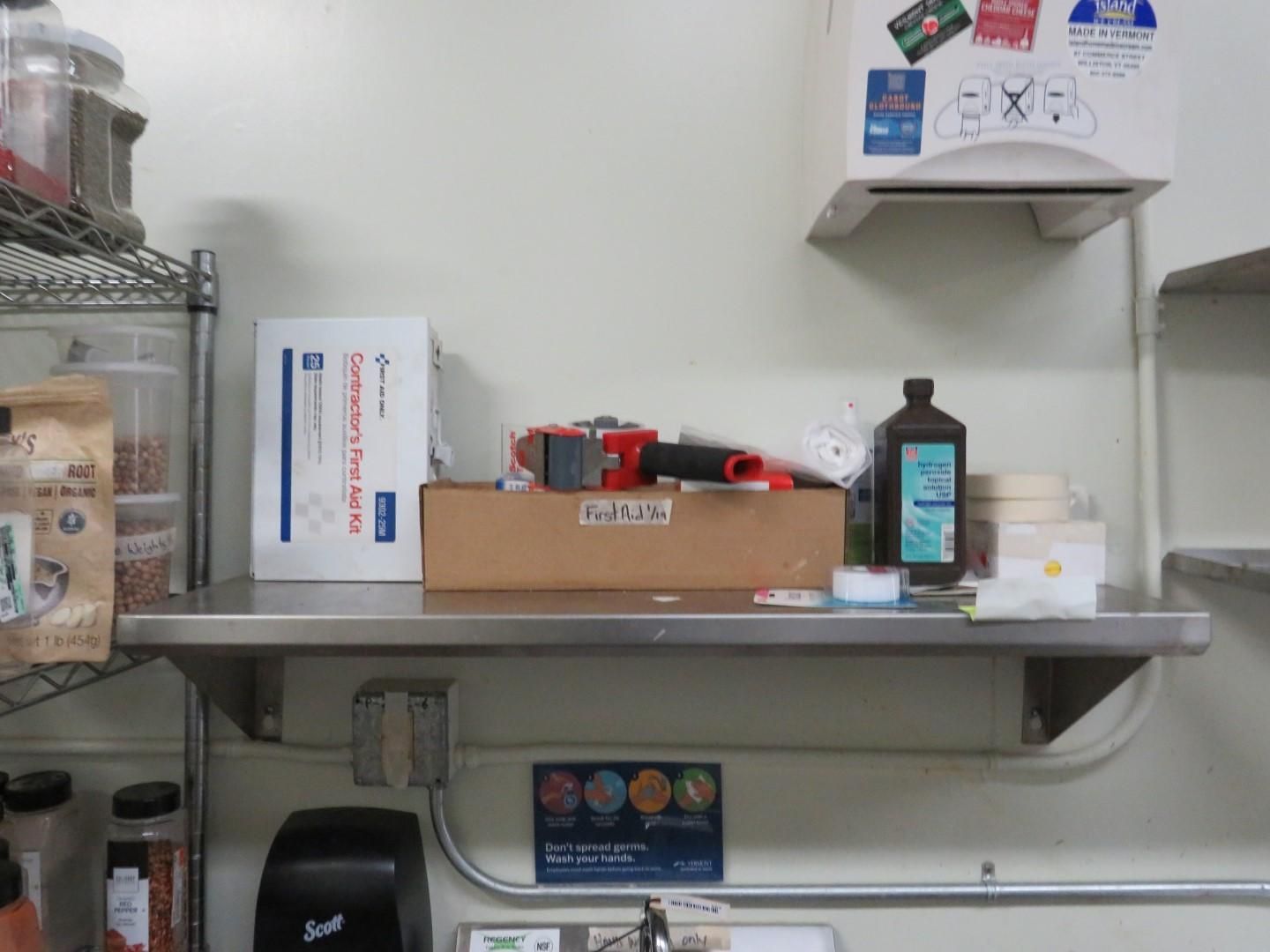 A stainless steel shelf in a kitchen holding boxes, bottles, and a paper towel dispenser against a white wall.