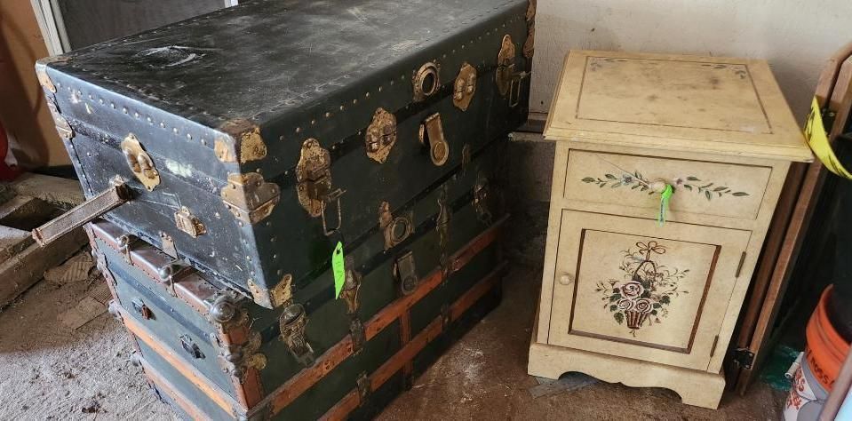 Two antique black trunks stacked; a painted cream nightstand is beside the trunks.