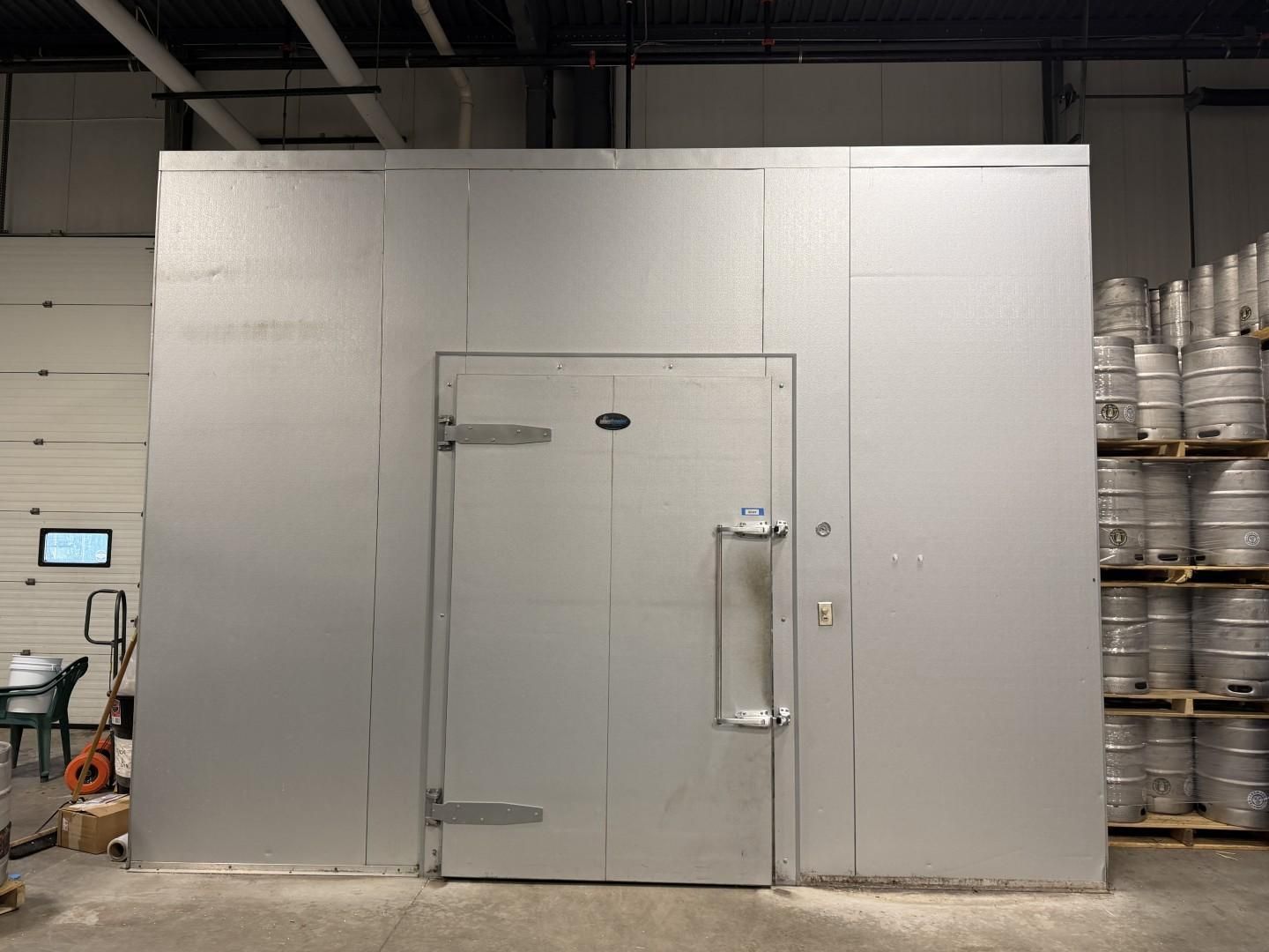 Metal walk-in cooler with door, handle, and hinges. Inside a warehouse, kegs are stacked to the right.