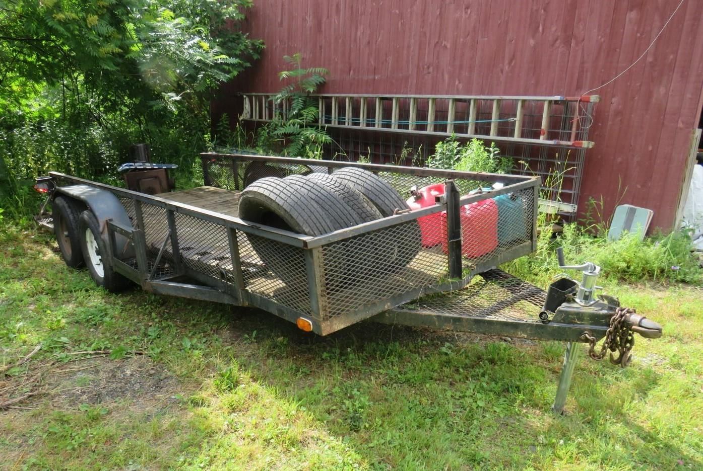 Black utility trailer with tires and containers, parked on grass near a red barn and trees.