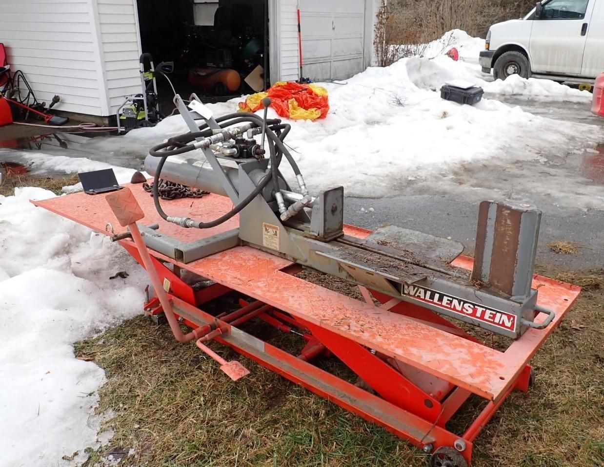 A gray wood splitter mounted on an orange hydraulic lift table in a snowy yard with a garage in the background.