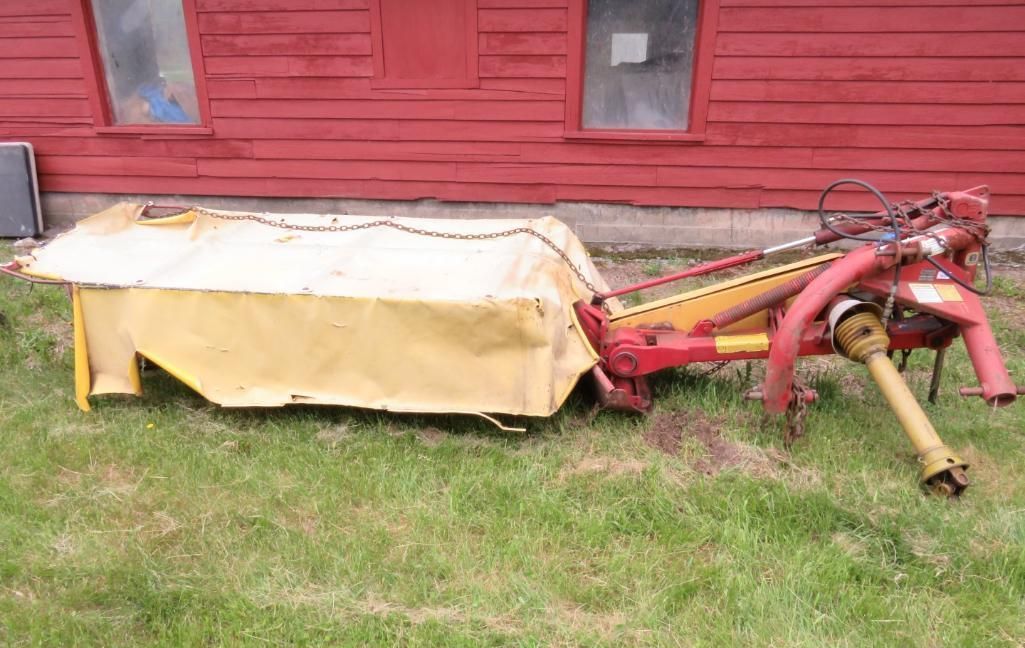 A yellow and red hay mower on grass in front of a red building.