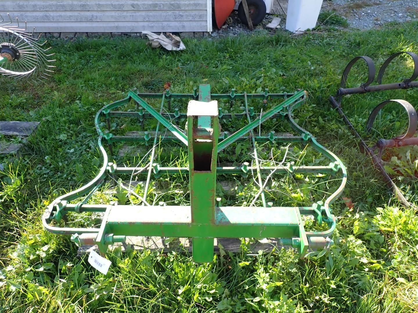 Green metal farm implement on grass beside a building, with curved tines and a central frame.