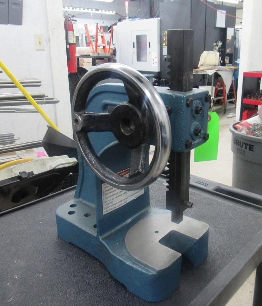 Blue metal press machine with a handwheel, on a dark table, in a workshop.
