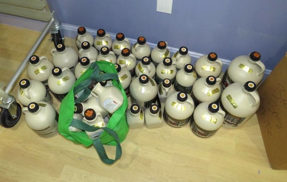 Bottles of maple syrup, mostly on a floor. A green reusable shopping bag holds more.