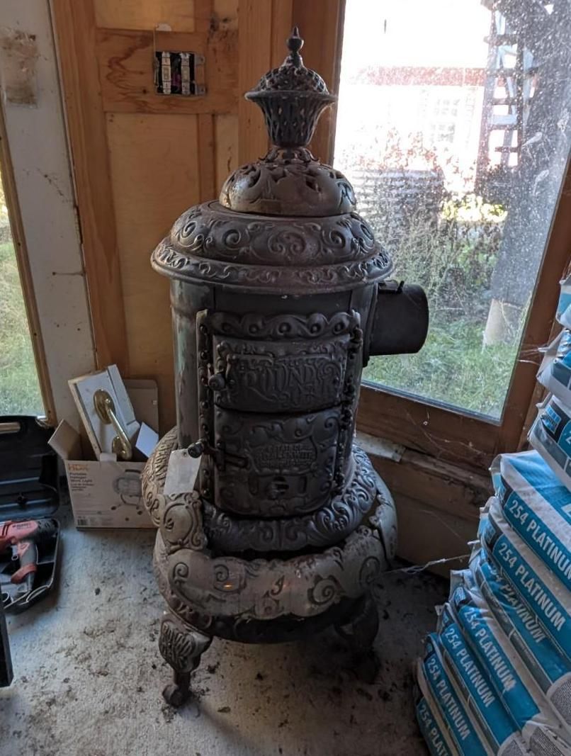 Old cast-iron stove in a sunlit wooden room beside a window and stacked blue-packaged items