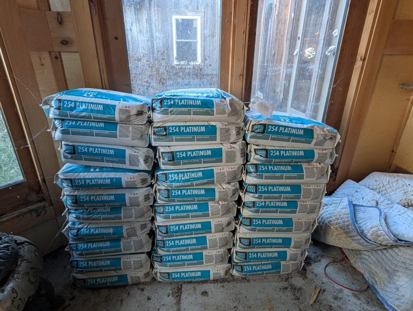 Stacked blue-and-white bags of building mix in a wooden room, beside a folded blanket and window.