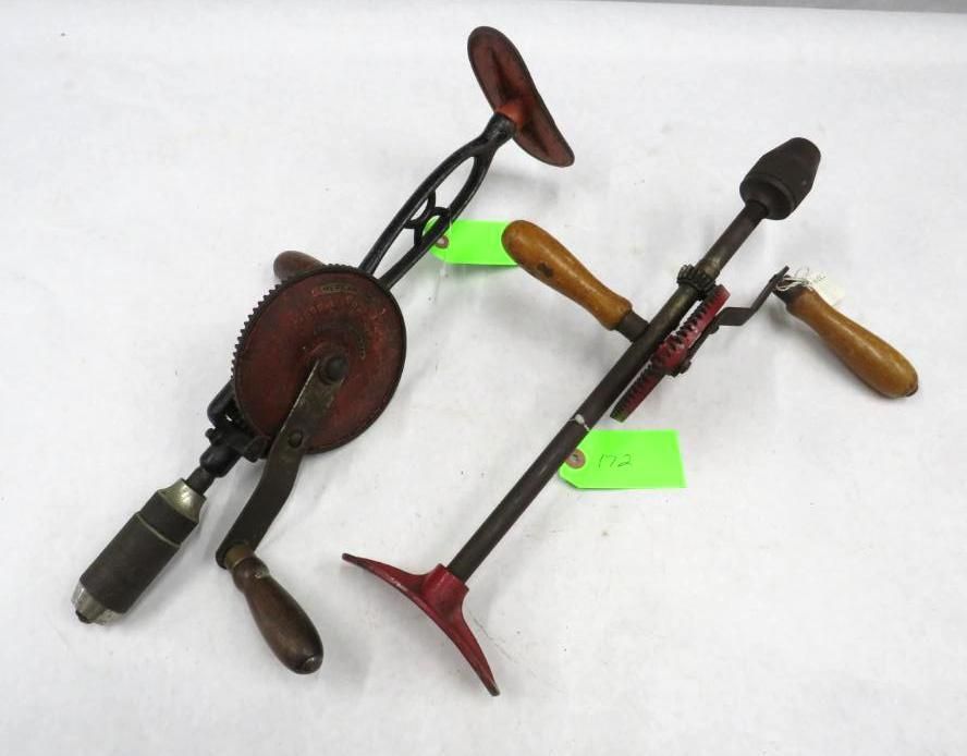 Two antique hand drills with wooden handles and metal frames on a white surface.