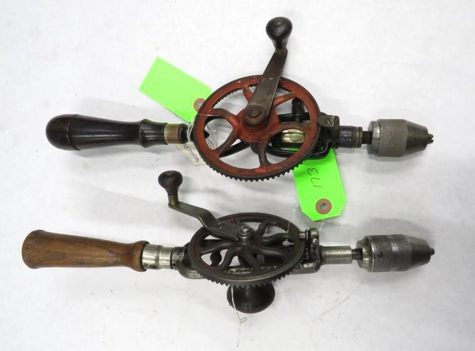Two antique hand-cranked drill presses on a white surface, one with a red gear, the other with a dark gear.