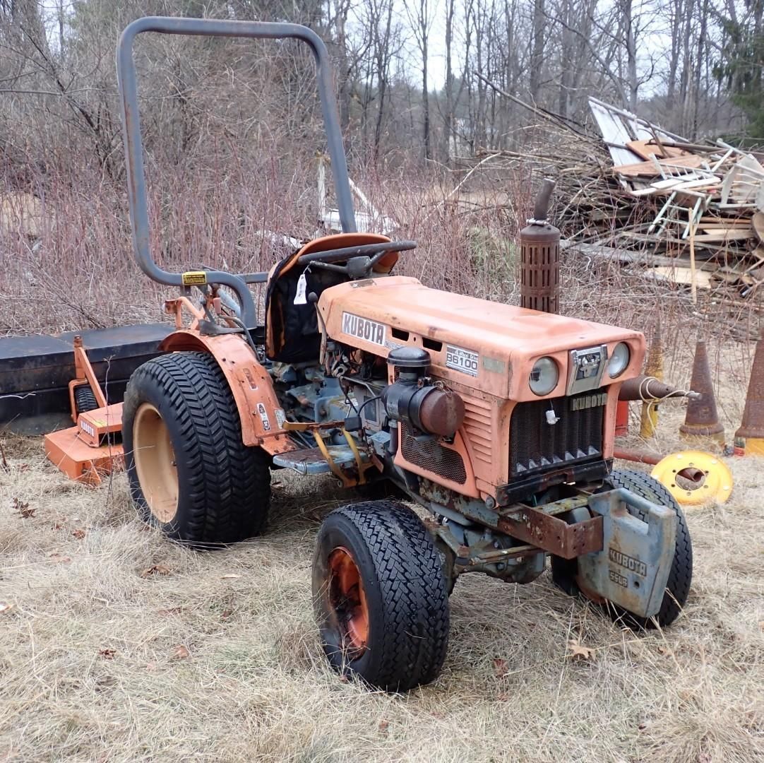 An orange compact utility tractor sits outdoors in a field with a mower attachment on the back and a brush guard.