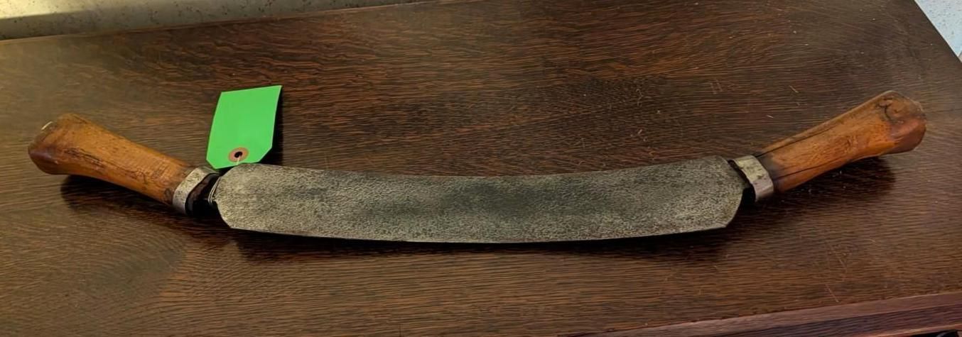 A vintage drawknife with wooden handles and a curved metal blade, resting on a dark wood surface with a green paper tag.