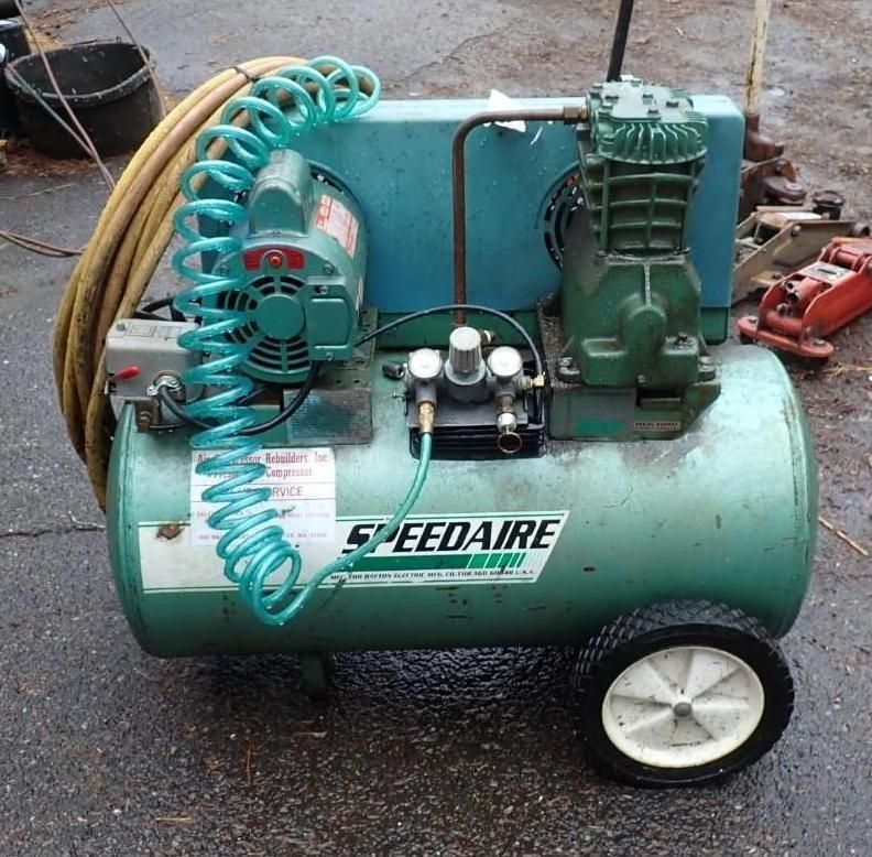 A teal Speedaire air compressor on wheels with a coiled blue hose, sitting on a paved surface.