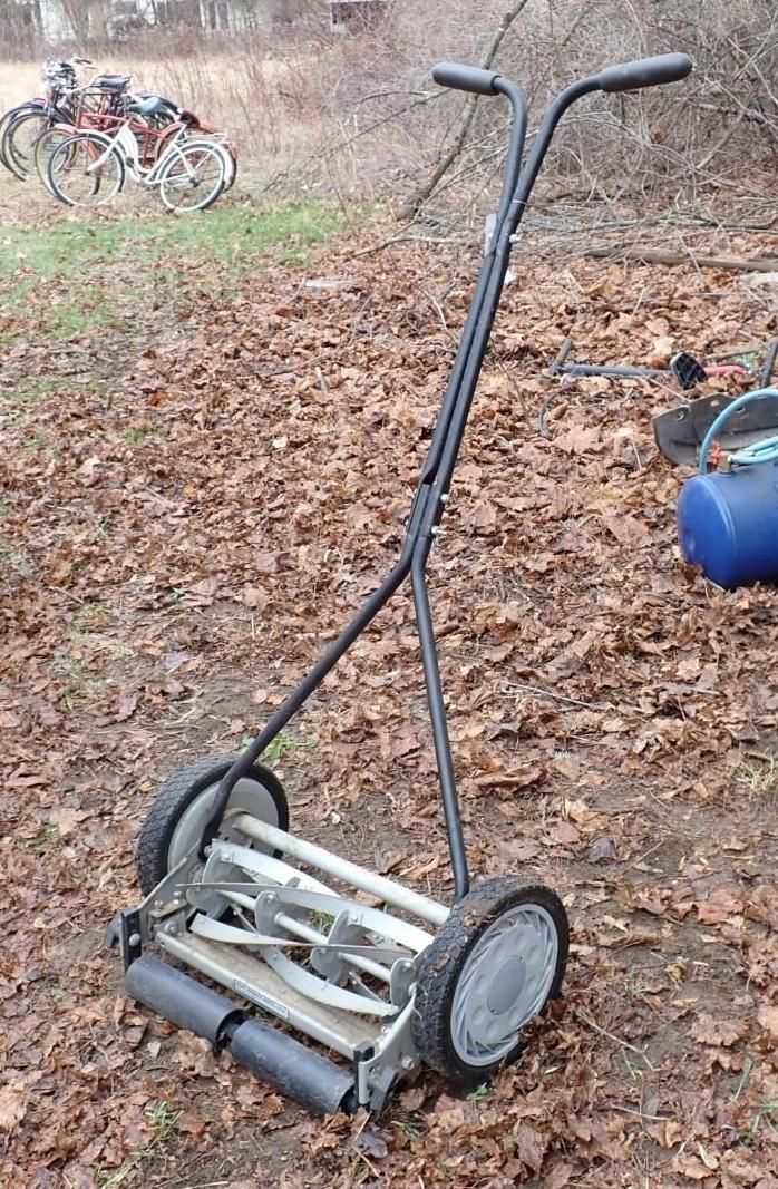 A manual reel lawnmower with a black handle and grey wheels sits on a lawn.