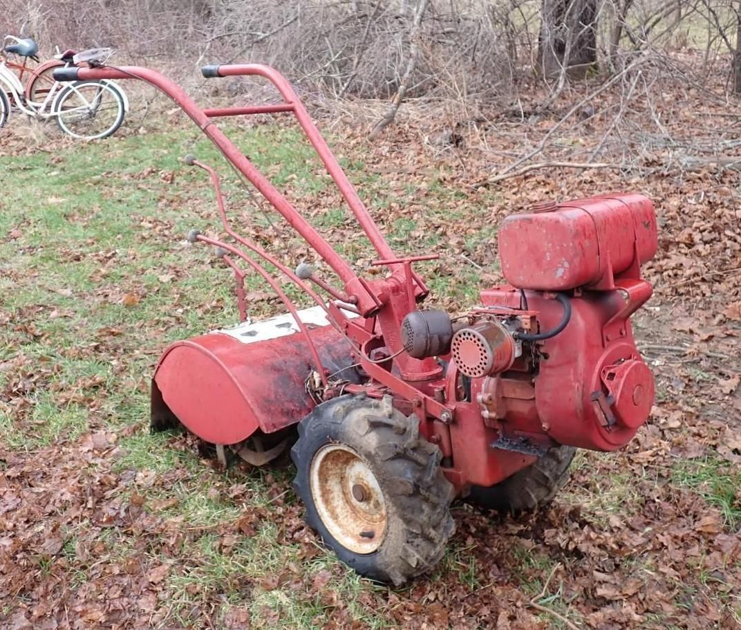 A vintage red Troy-Bilt rototiller with knobby tires sits on a lawn.
