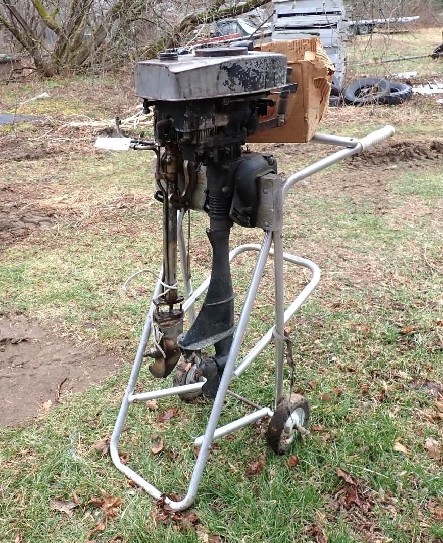 An outboard motor mounted on a metal wheeled stand, sitting in a grassy, outdoor area.