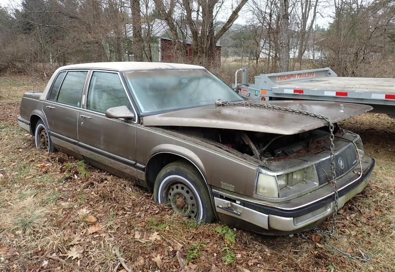 A brown vintage sedan sits in a field, a metal chain sits across its open hood.