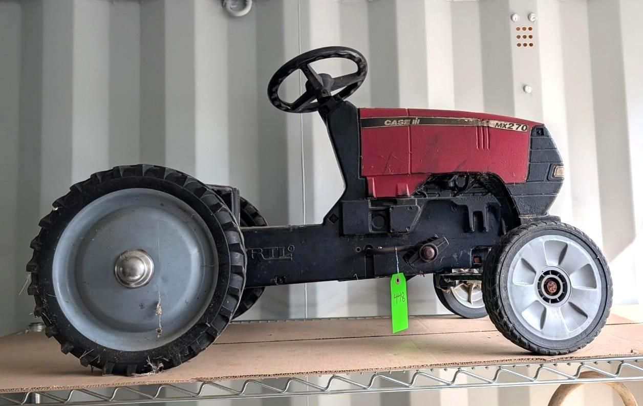 A red and black toy tractor with large gray wheels, sitting on a shelf against a corrugated metal wall.