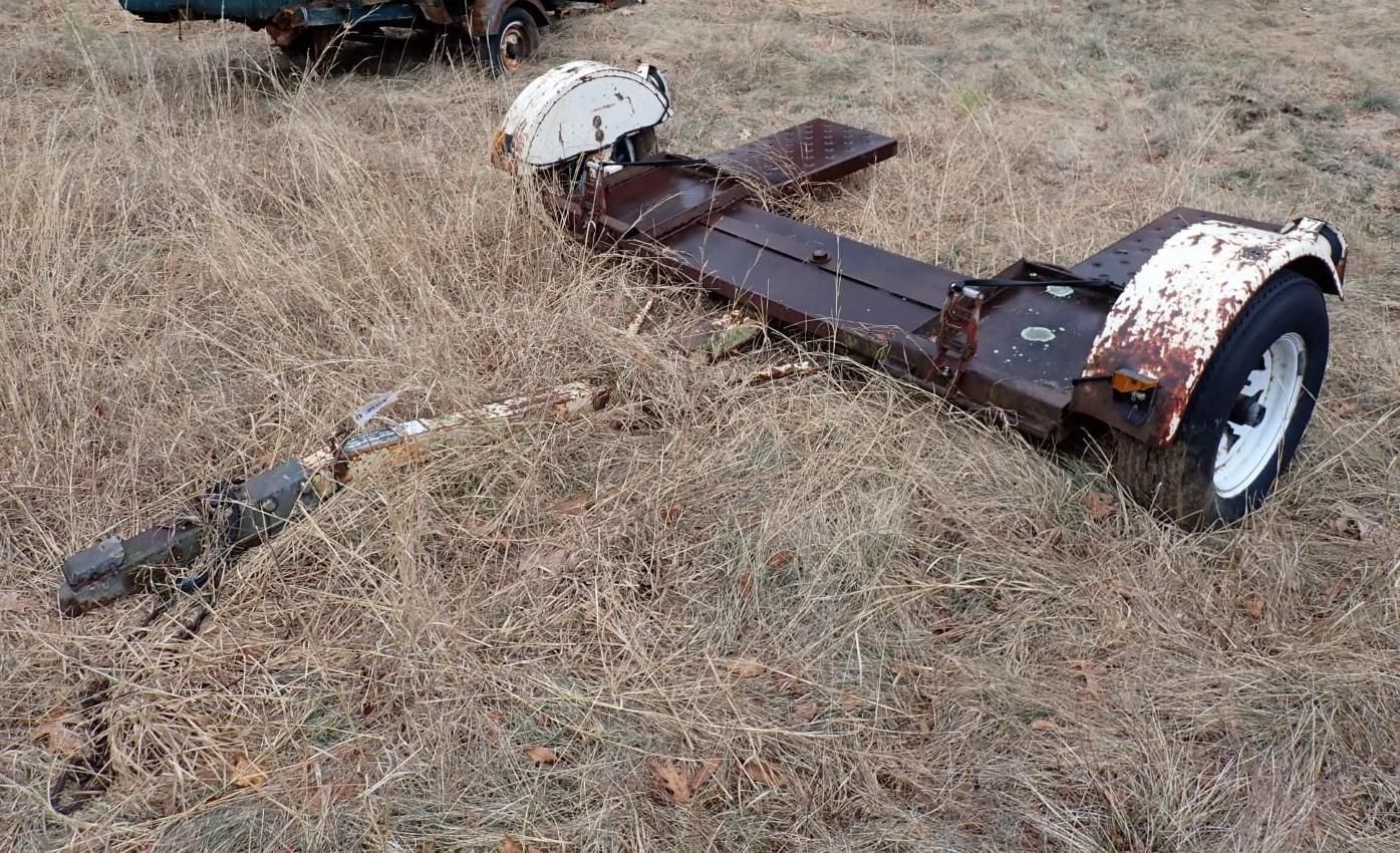 A metal car tow dolly sits in a grassy field with one visible wheel and a white fender.