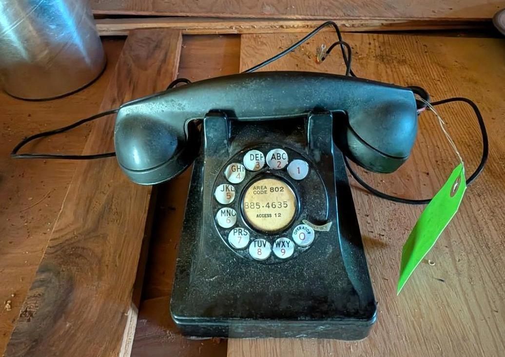 A vintage, black rotary telephone sits on a wooden surface, with a small green price tag attached to its cord.