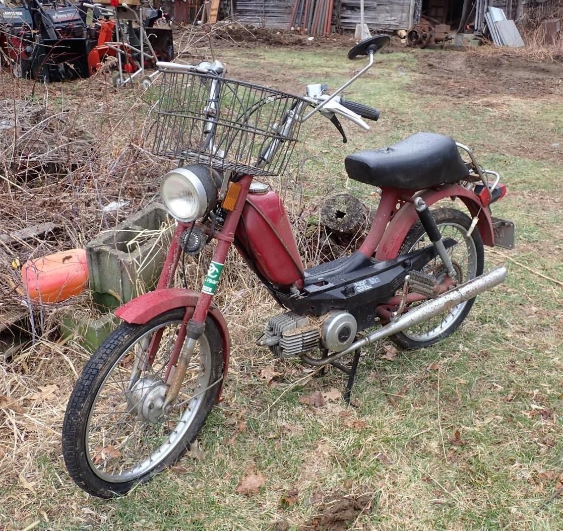 A red vintage moped with a wire basket sits on a grassy, outdoor lot.