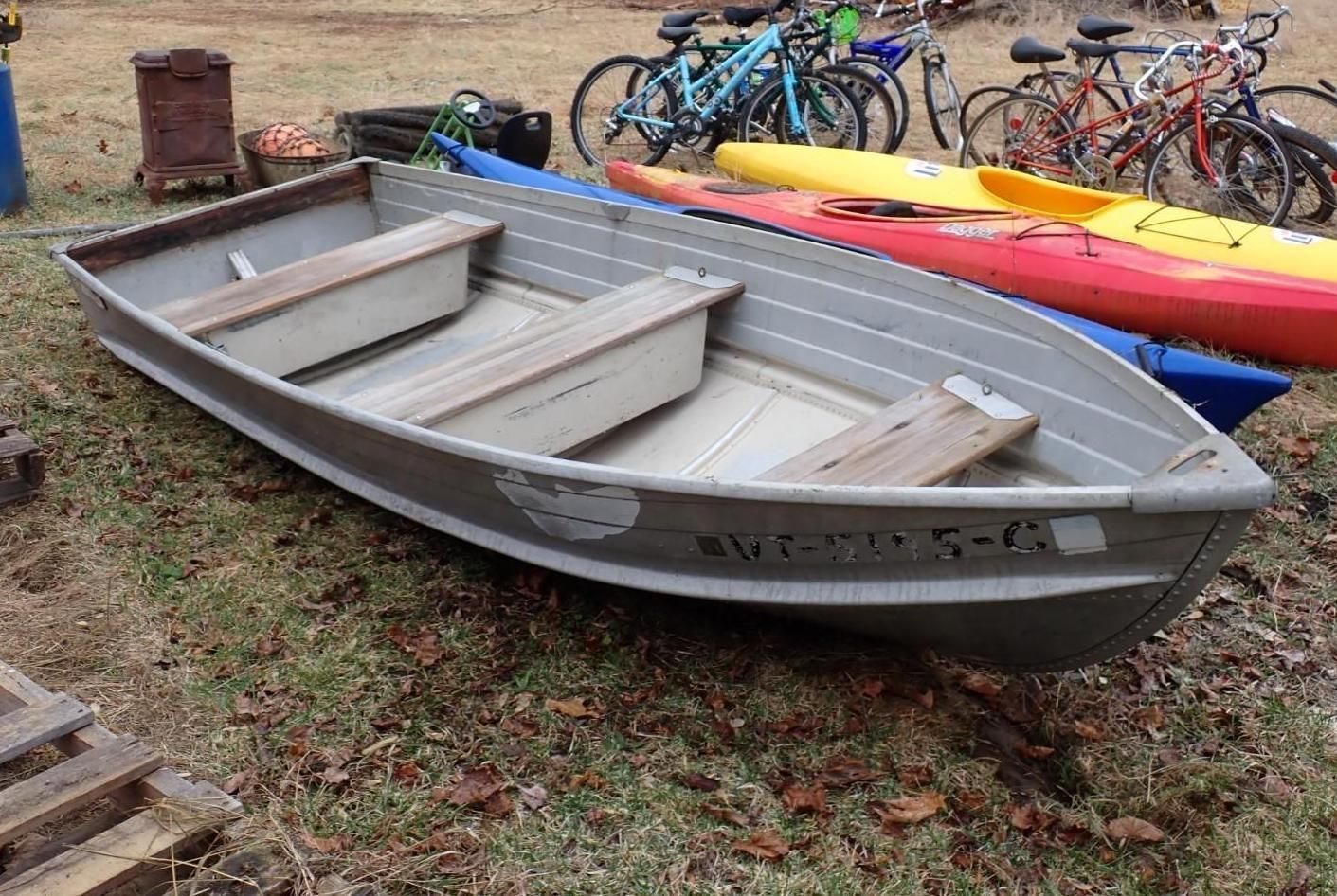 A gray aluminum utility boat sits on a grass lawn next to a red and a yellow kayak and several bicycles.