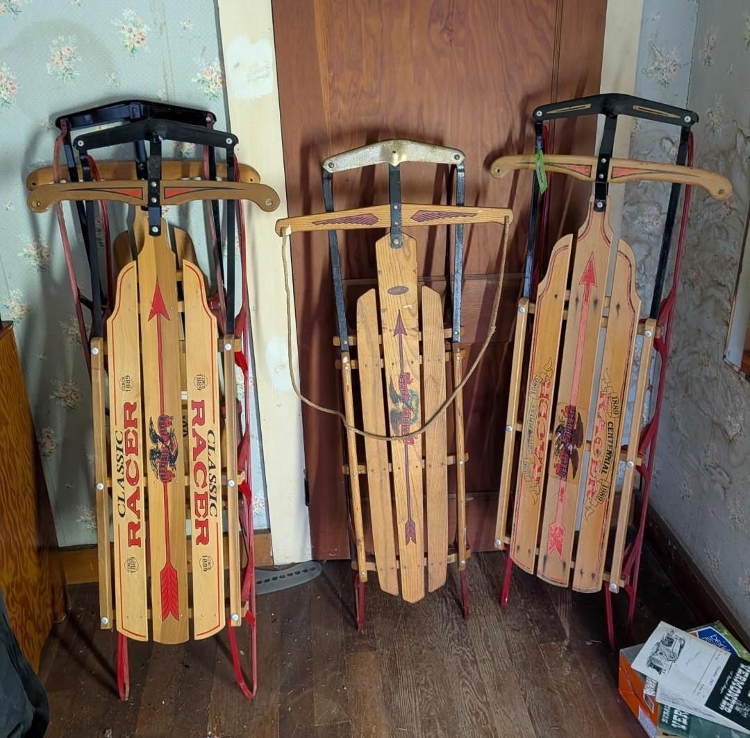 Three vintage wooden sleds with metal runners standing upright against a wooden door and patterned wallpaper.