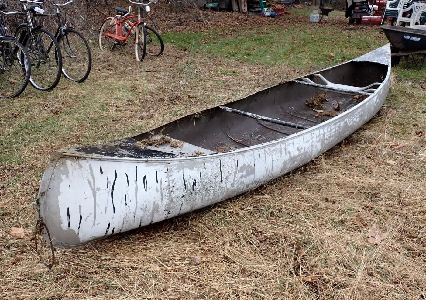 A white aluminum canoe rests on the grass near two bicycles in a yard.
